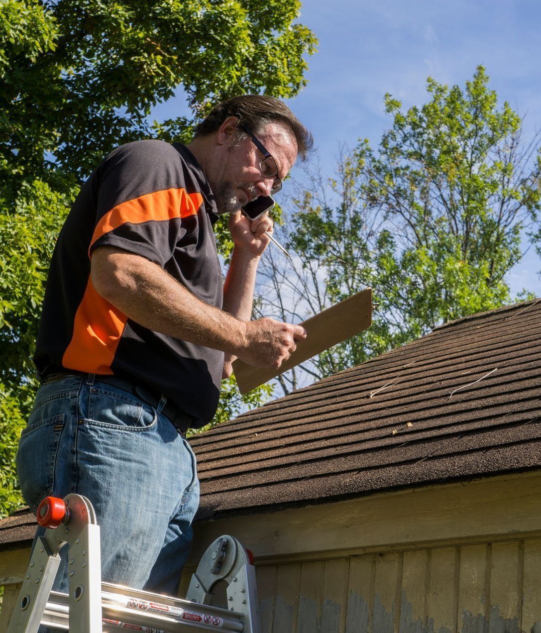 Worker on a ladder inspecting a roof while talking on a phone, holding a clipboard.