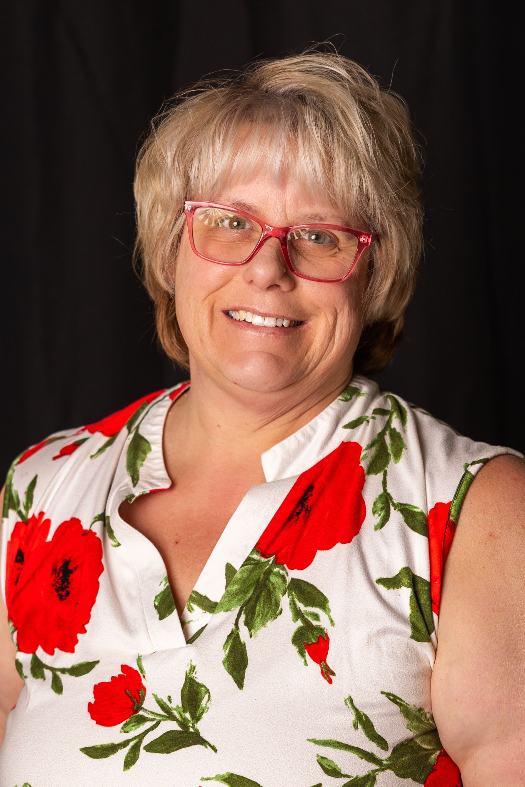 Woman with blond hair and red-framed glasses smiles, wearing a white top with red floral print; black background.