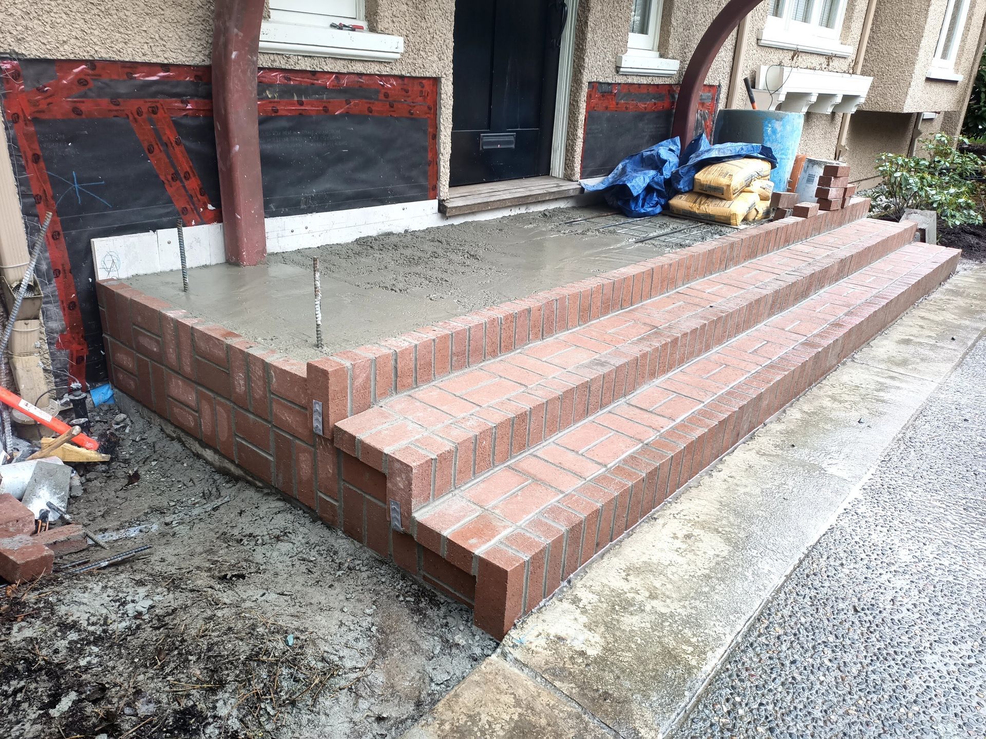 Brick front steps and porch under construction, with wet concrete and tools nearby.