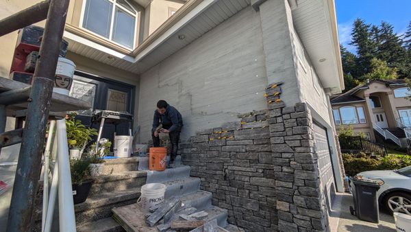 Person working on stone steps beside a house, with buckets and stacked materials.