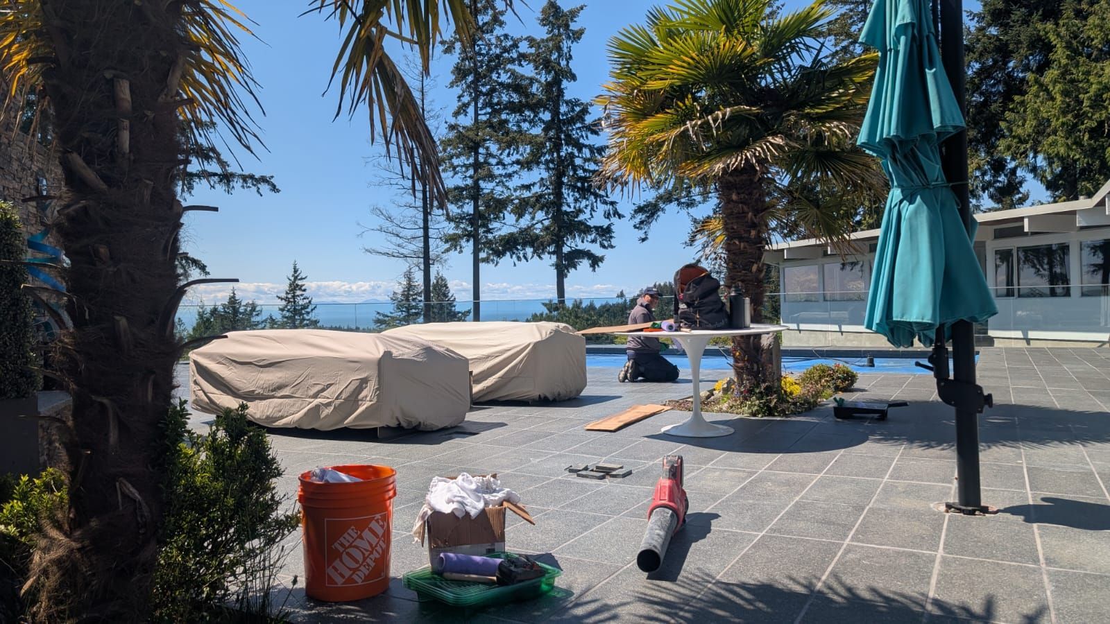 Patio by a lake with covered lounge chairs, trees, umbrellas, and cleaning items in the foreground