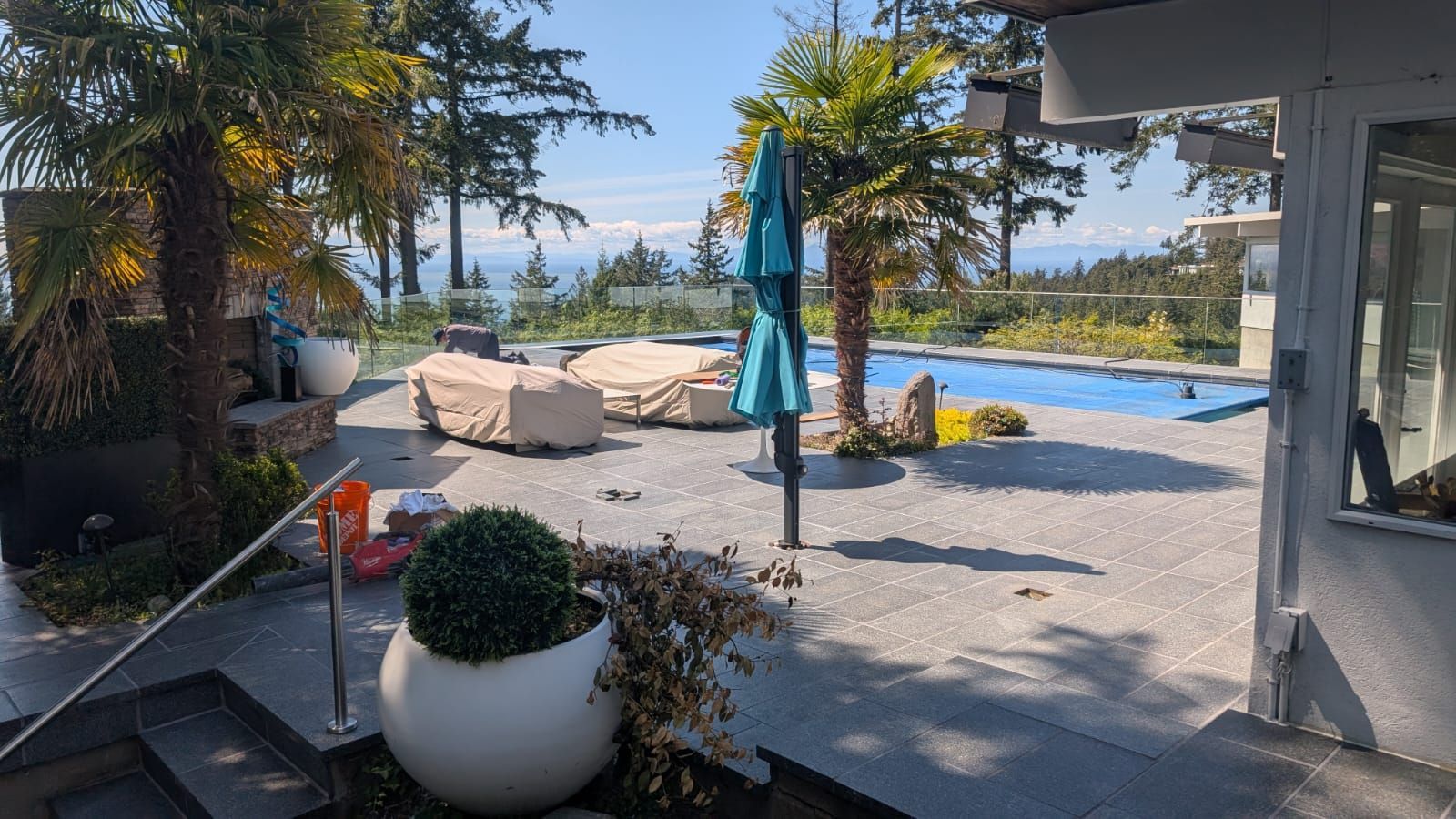 Sunny patio with lounge chairs, blue umbrella, potted plant, and poolside view of the landscape