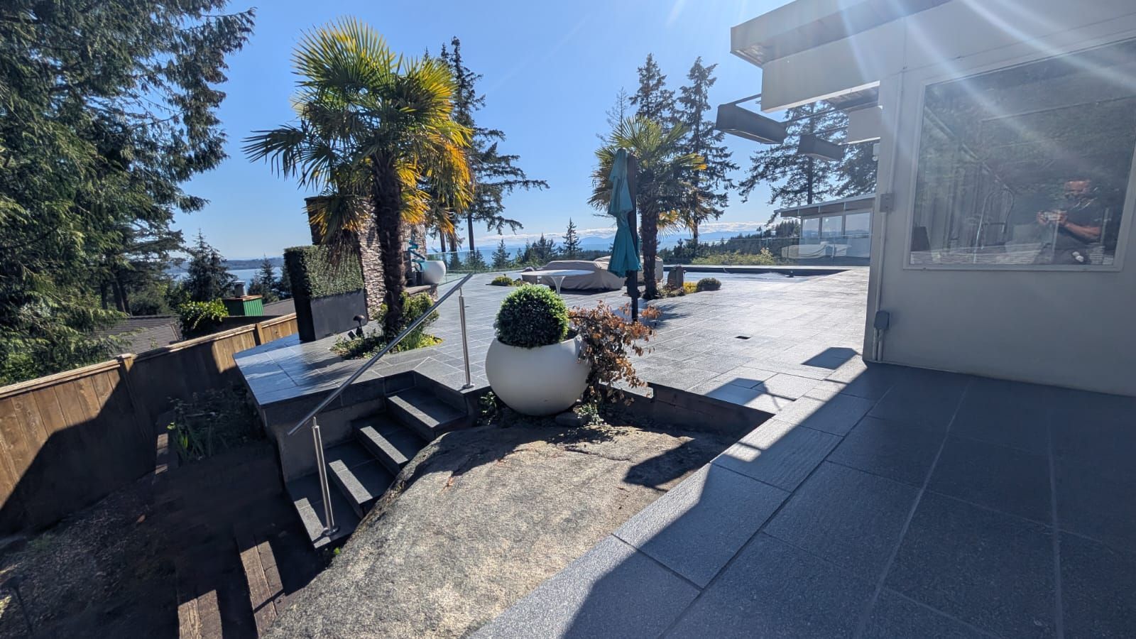 Sunny patio with potted plants, trees, and outdoor seating beside a building