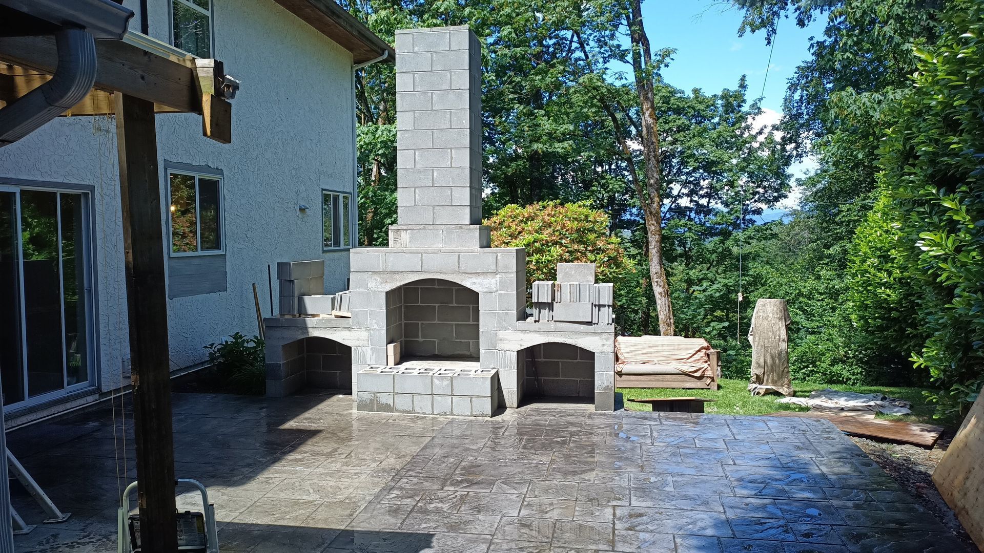 Outdoor stone pizza oven and fireplace on a patio beside a house, with trees and a wet paved ground.