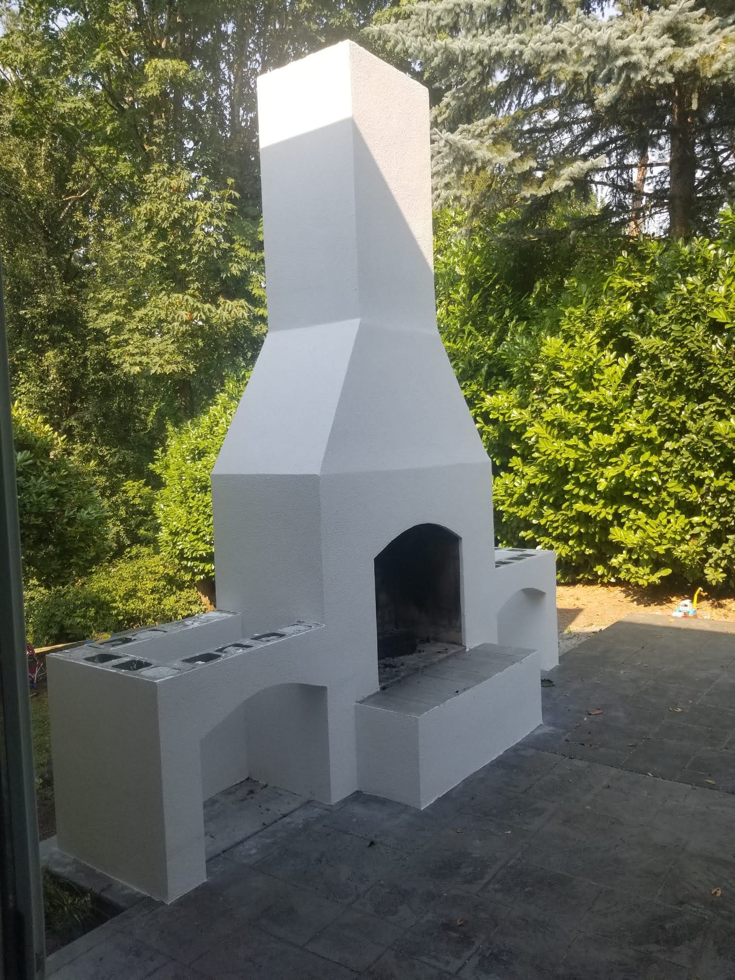 Outdoor white stucco fireplace with bench in a garden, surrounded by greenery and trees
