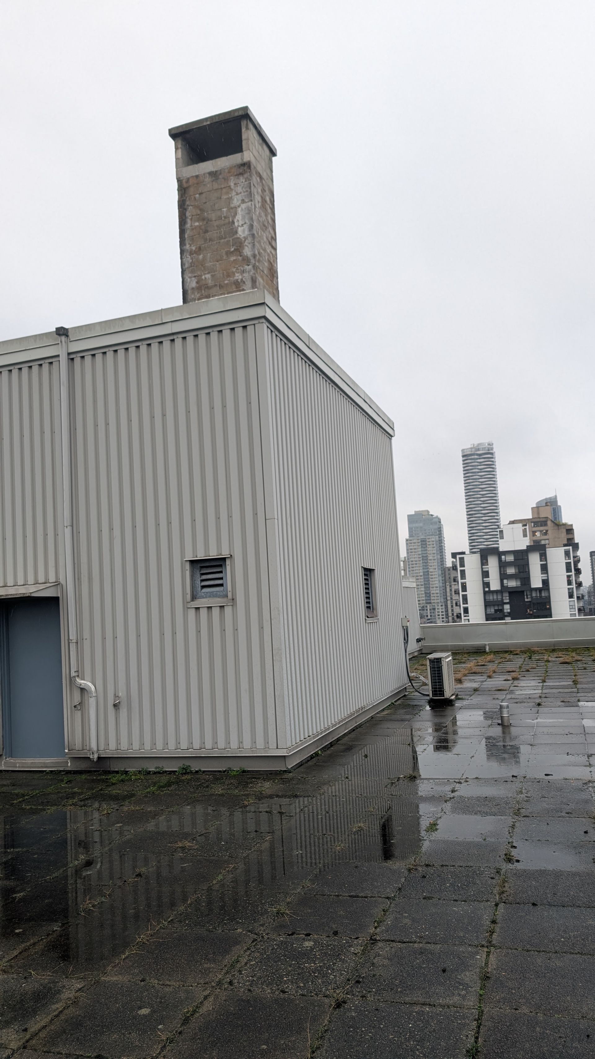 Rooftop view of a gray corrugated building and chimney on a wet, overcast day with city buildings in the distance