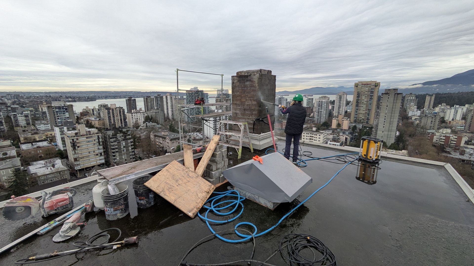 Worker on a rooftop with tools and cables, overlooking a dense city skyline at sunset.