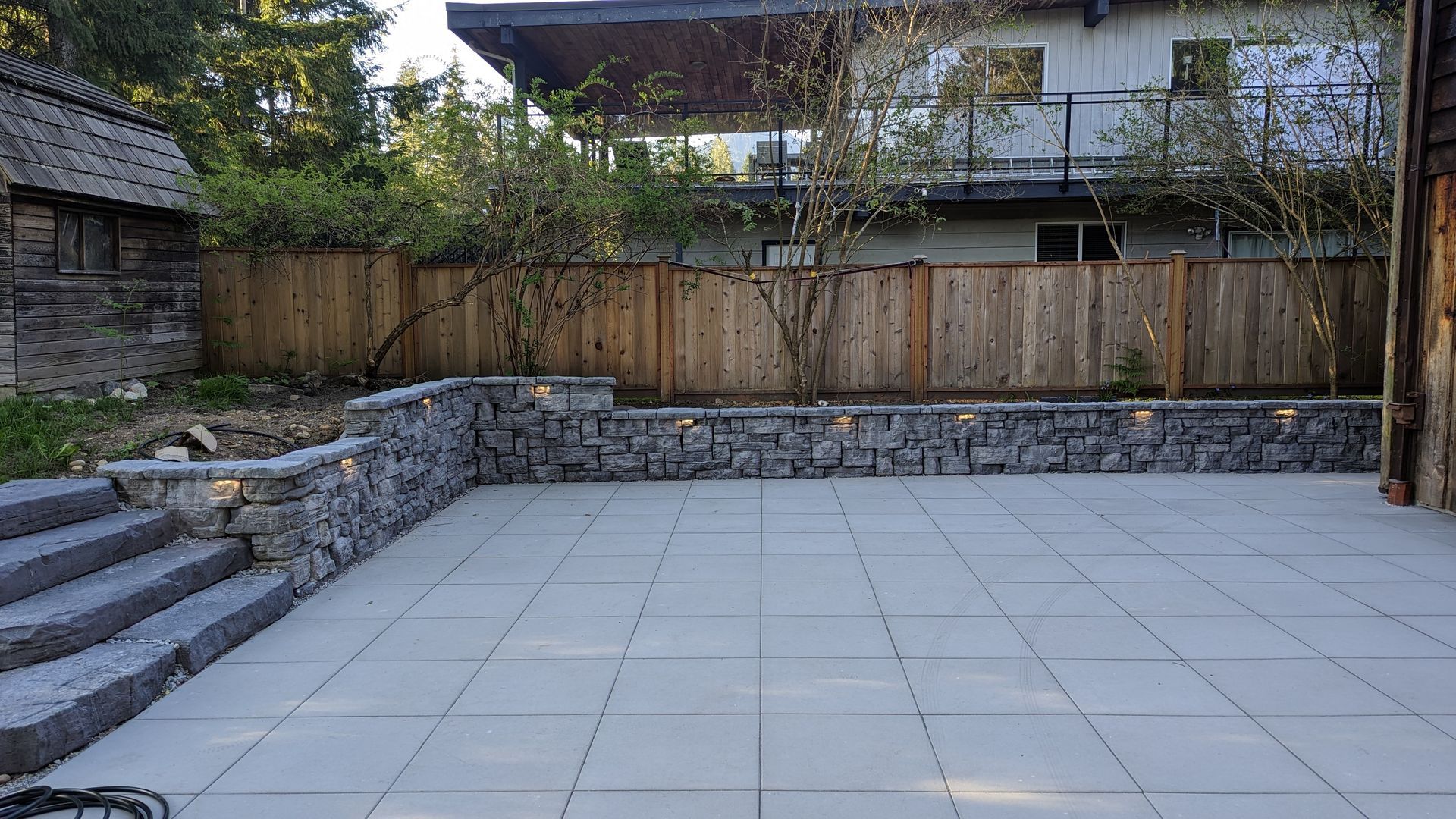 A concrete slab patio surrounded by a short natural stone retaining wall in the back yard of a residential house in Vancouver, Canada.