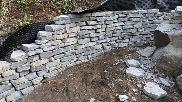 Curved stacked-stone retaining wall beside a dirt slope and scattered rocks outdoors