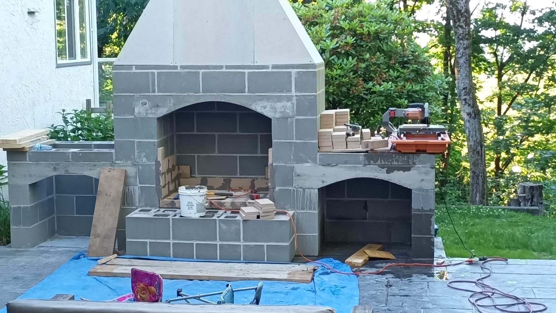 Outdoor brick fireplace under construction with tools and materials on a blue tarp in a yard