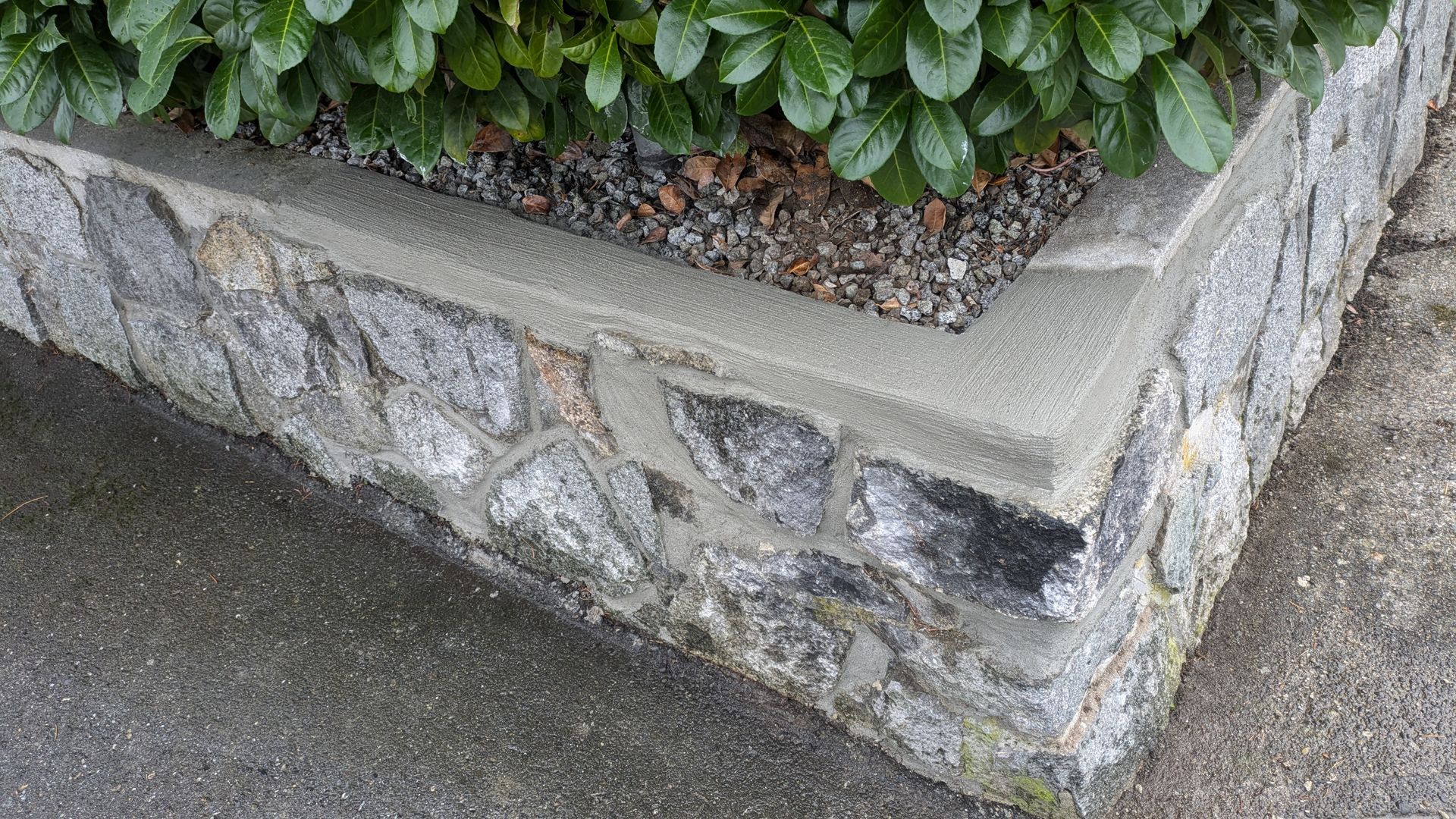 Stone planter wall with concrete cap and green shrubs beside a paved sidewalk.