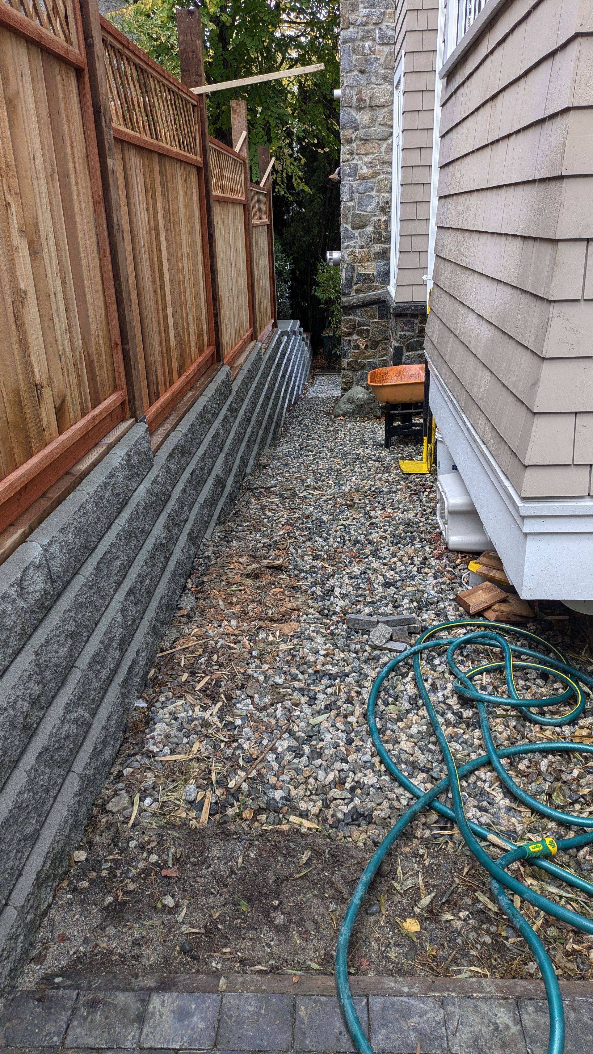 Narrow gravel side yard beside a house, with a wooden fence, gray siding, and a coiled green hose.