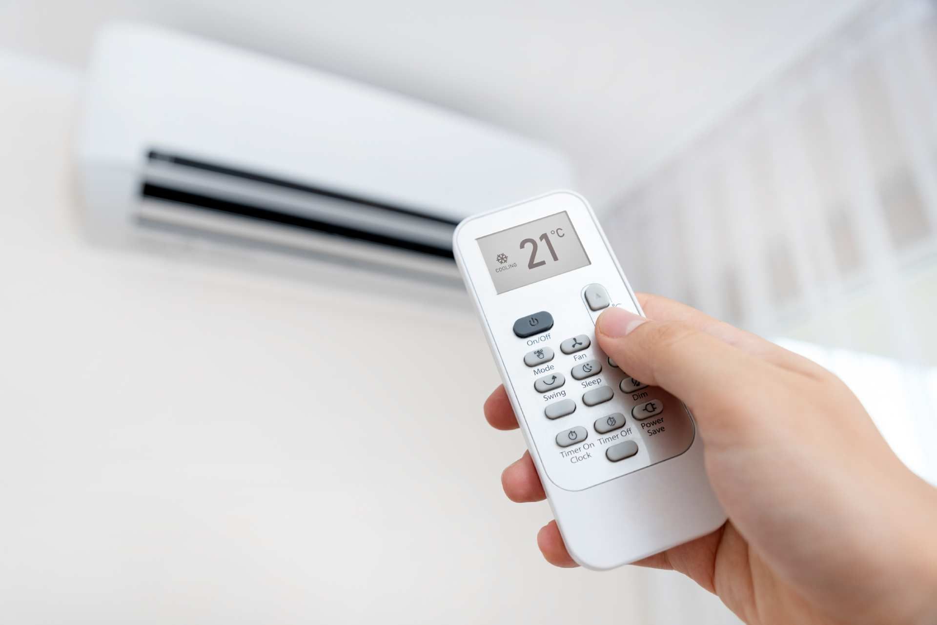 Hand holding a remote pointed at a wall-mounted air conditioner in a bright room
