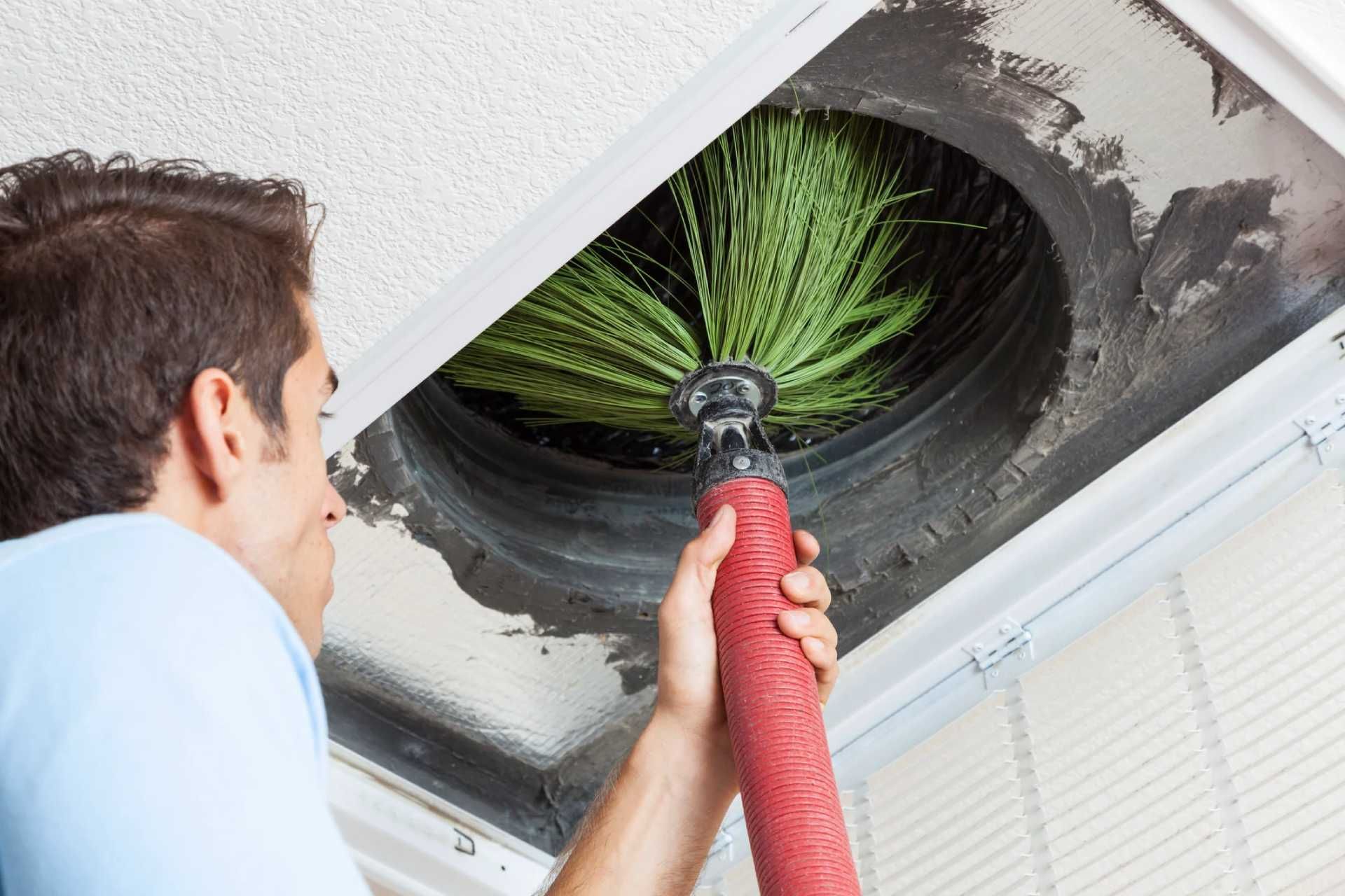 Worker cleaning a ceiling vent with a red vacuum hose, exposing green dust inside the duct