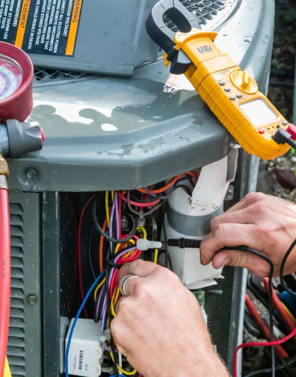 Hands wiring an outdoor HVAC unit with a yellow multimeter on top.