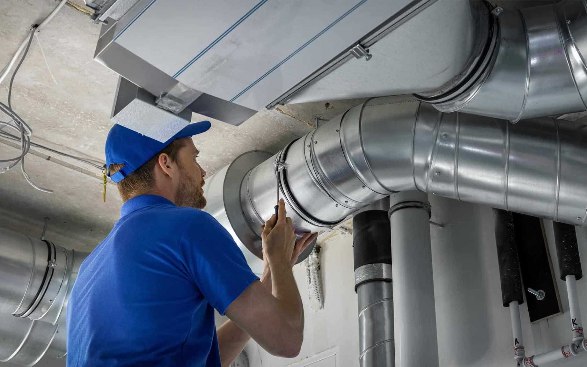 Technician in blue uniform inspecting metal ductwork in an industrial ceiling