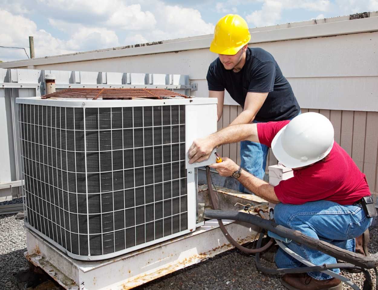 Two workers in hard hats repair an outdoor HVAC unit on a rooftop.
