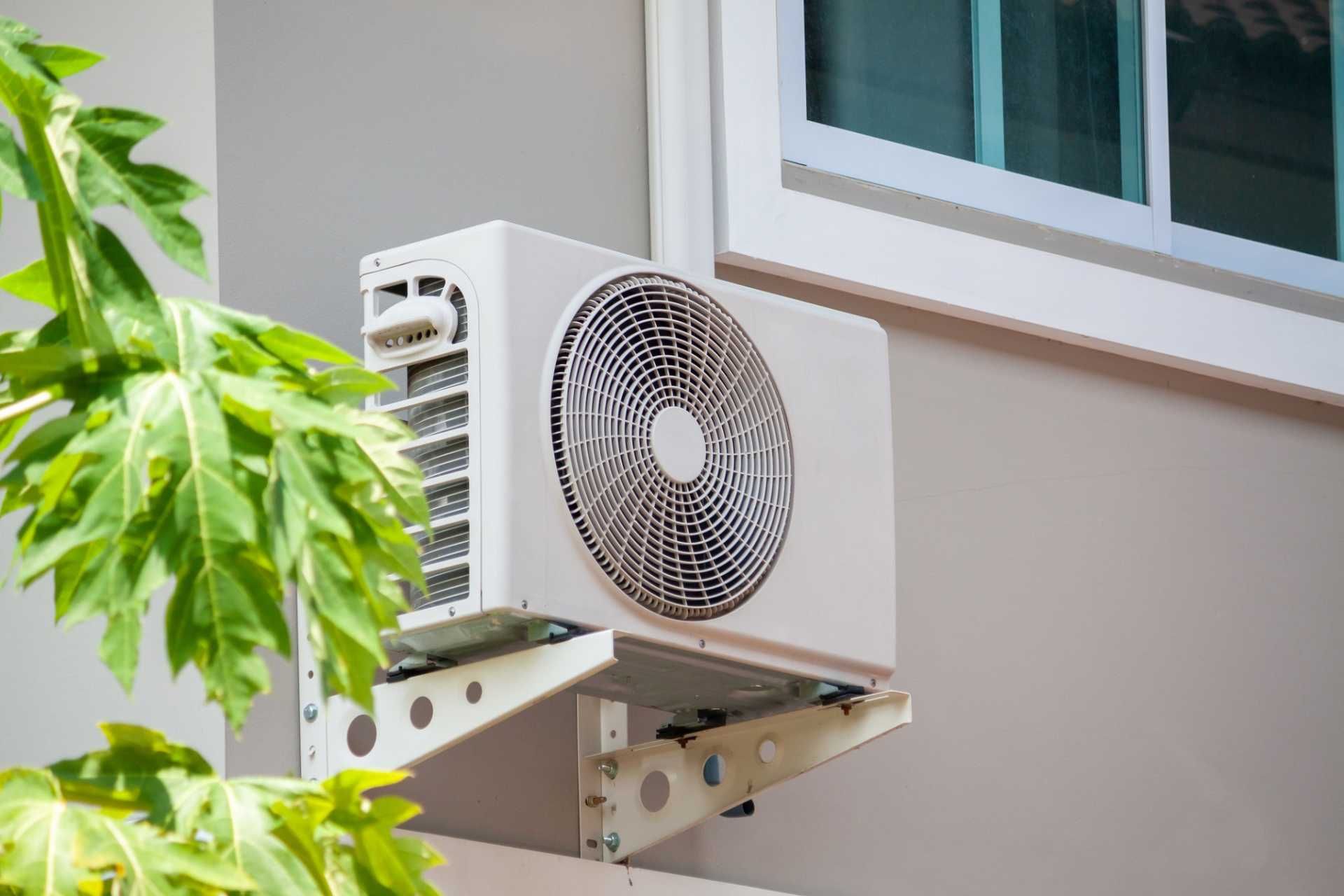 Outdoor wall-mounted air conditioner unit beside a building window, partially framed by green leaves.