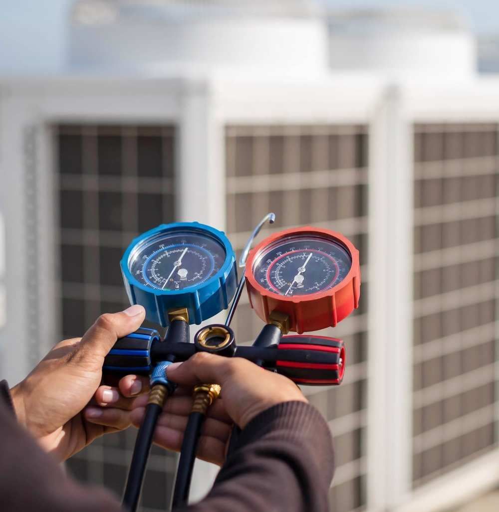 Hands holding blue and red HVAC pressure gauges in front of rooftop air conditioning units