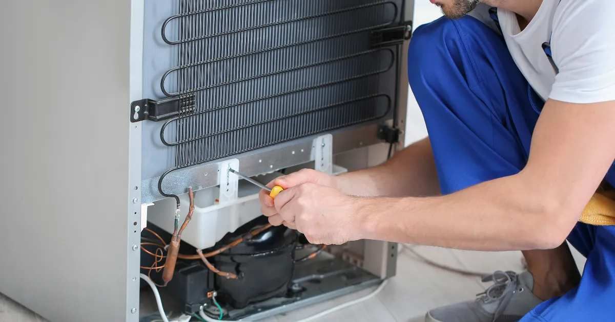 Technician in blue overalls repairing a refrigerator’s exposed compressor and coils