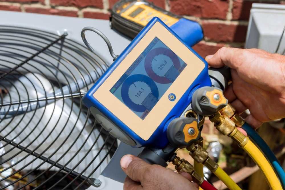 Hand holding HVAC manifold gauge set with blue screen beside outdoor air conditioner unit