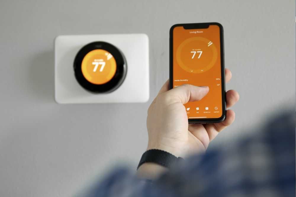 Person using a smartphone to control a wall-mounted thermostat showing 77°F