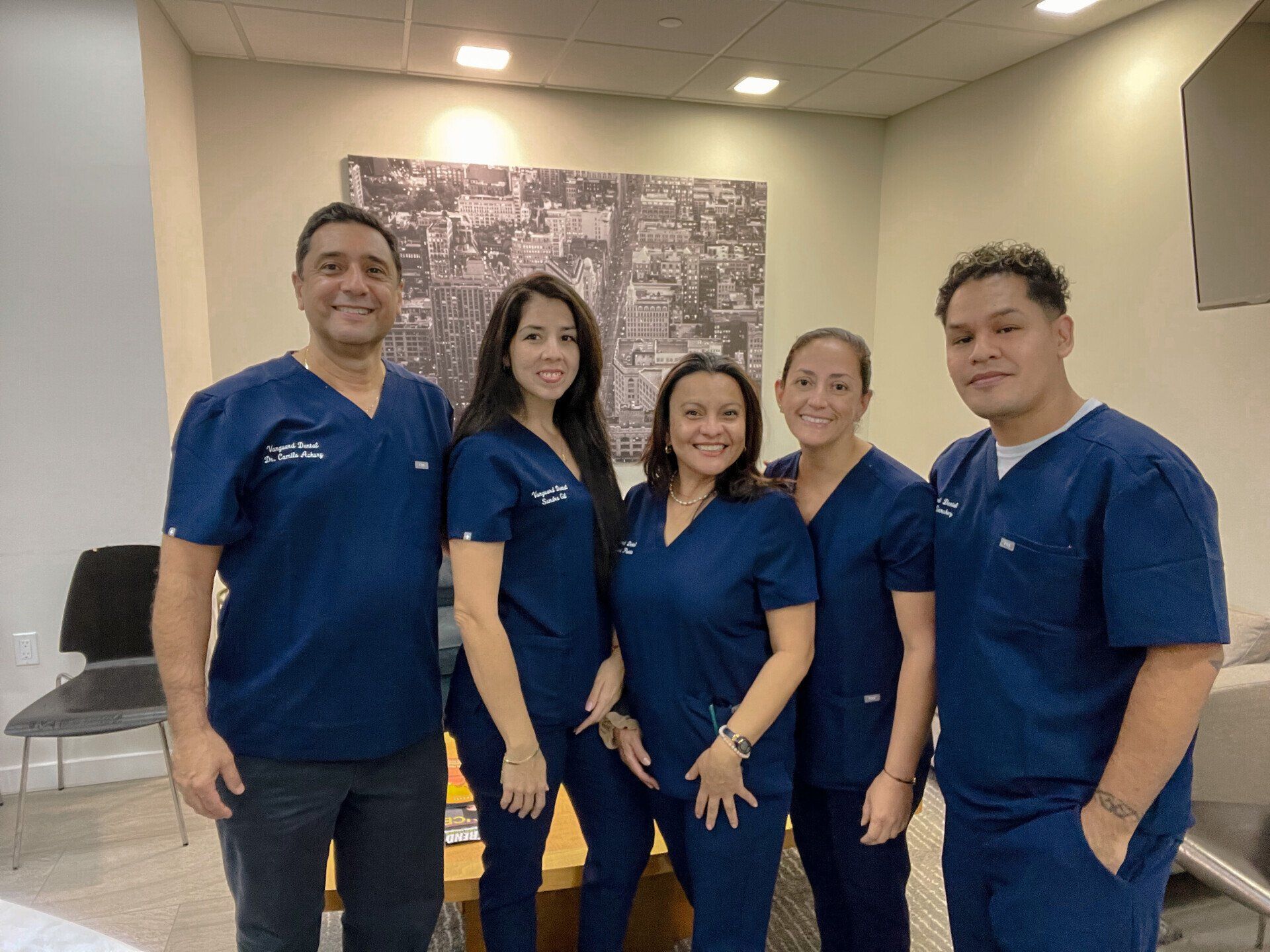 Five people in blue scrubs pose in a modern office, smiling in front of a neutral-toned wall with art.