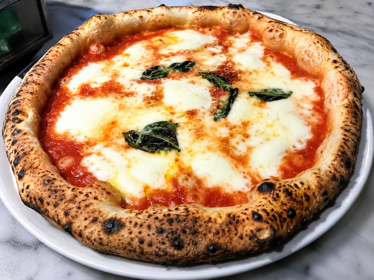 Pizza Margherita con foglie di basilico su un tavolo di legno, con una bevanda e un dolce sullo sfondo.