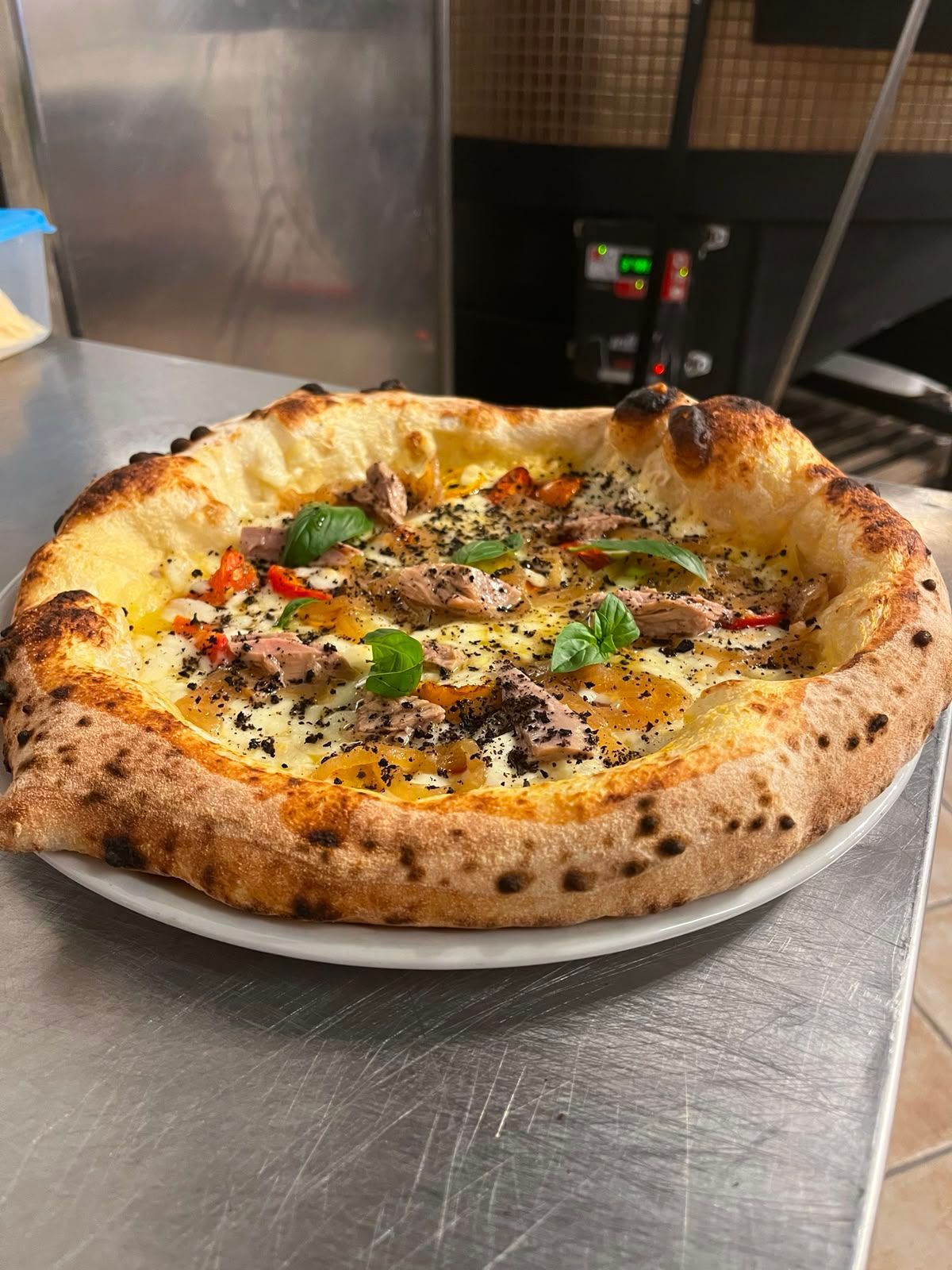 Pizza cotta nel forno a legna con formaggio fuso, basilico ed erbe aromatiche su un piano di lavoro in metallo.