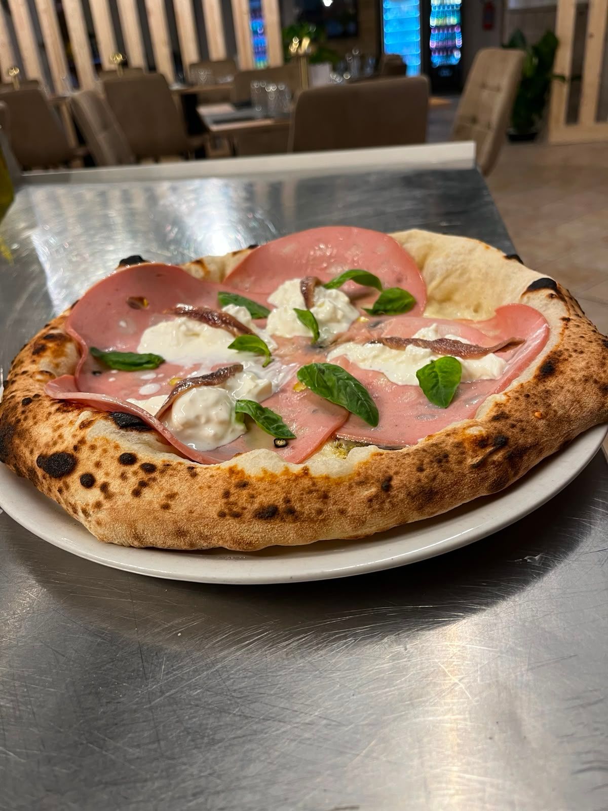 Una pizza a fette con prosciutto crudo, formaggio bianco e basilico, disposta su un piatto appoggiato su un tavolo di metallo.