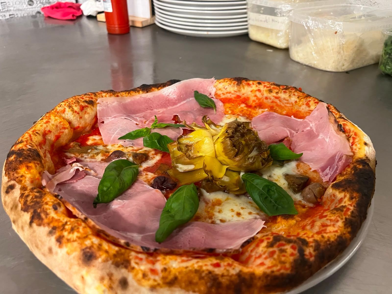 Primo piano di una pizza condita con prosciutto, basilico e verdure arrostite su un vassoio di metallo.