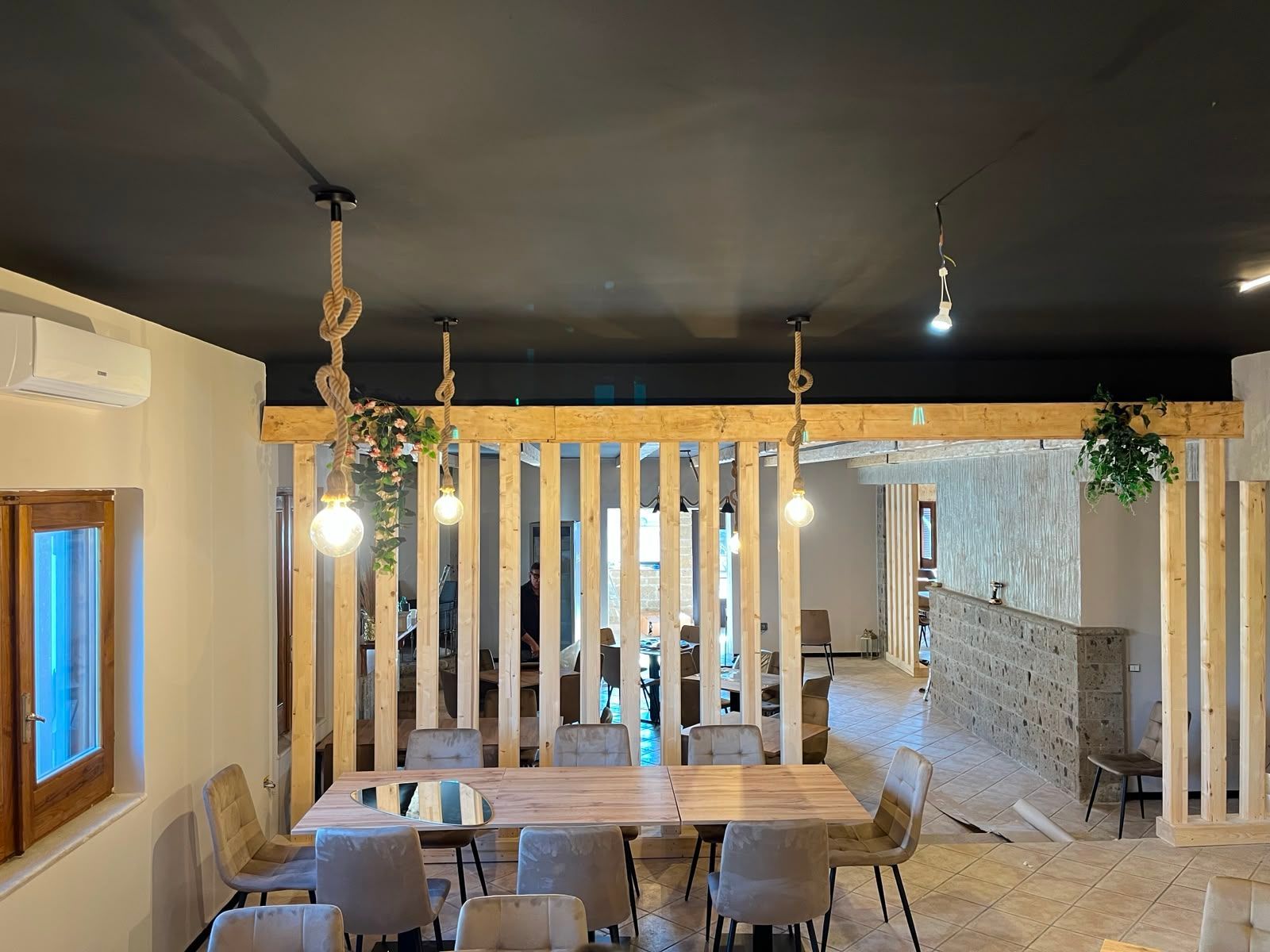 Accogliente zona pranzo con divisorio in legno, lampade a sospensione e un lungo tavolo accanto a una finestra e a un muro in pietra.