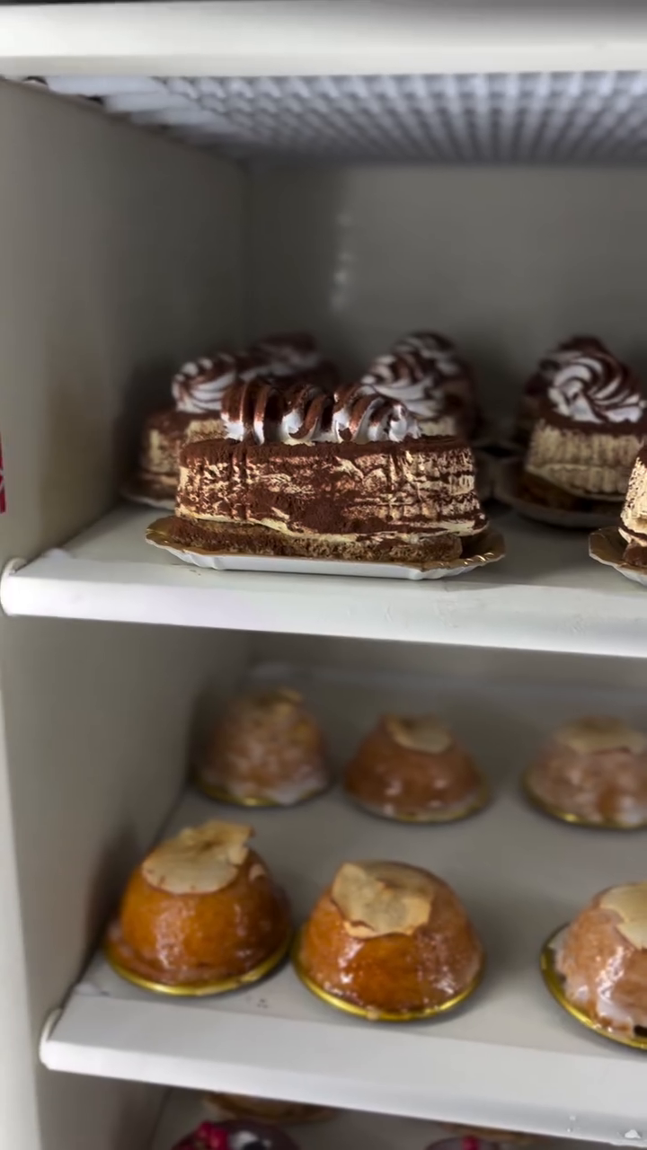 Pasticcini assortiti su ripiani bianchi del frigorifero, tra cui fette di torta ricoperte di cioccolato e brioche glassate.