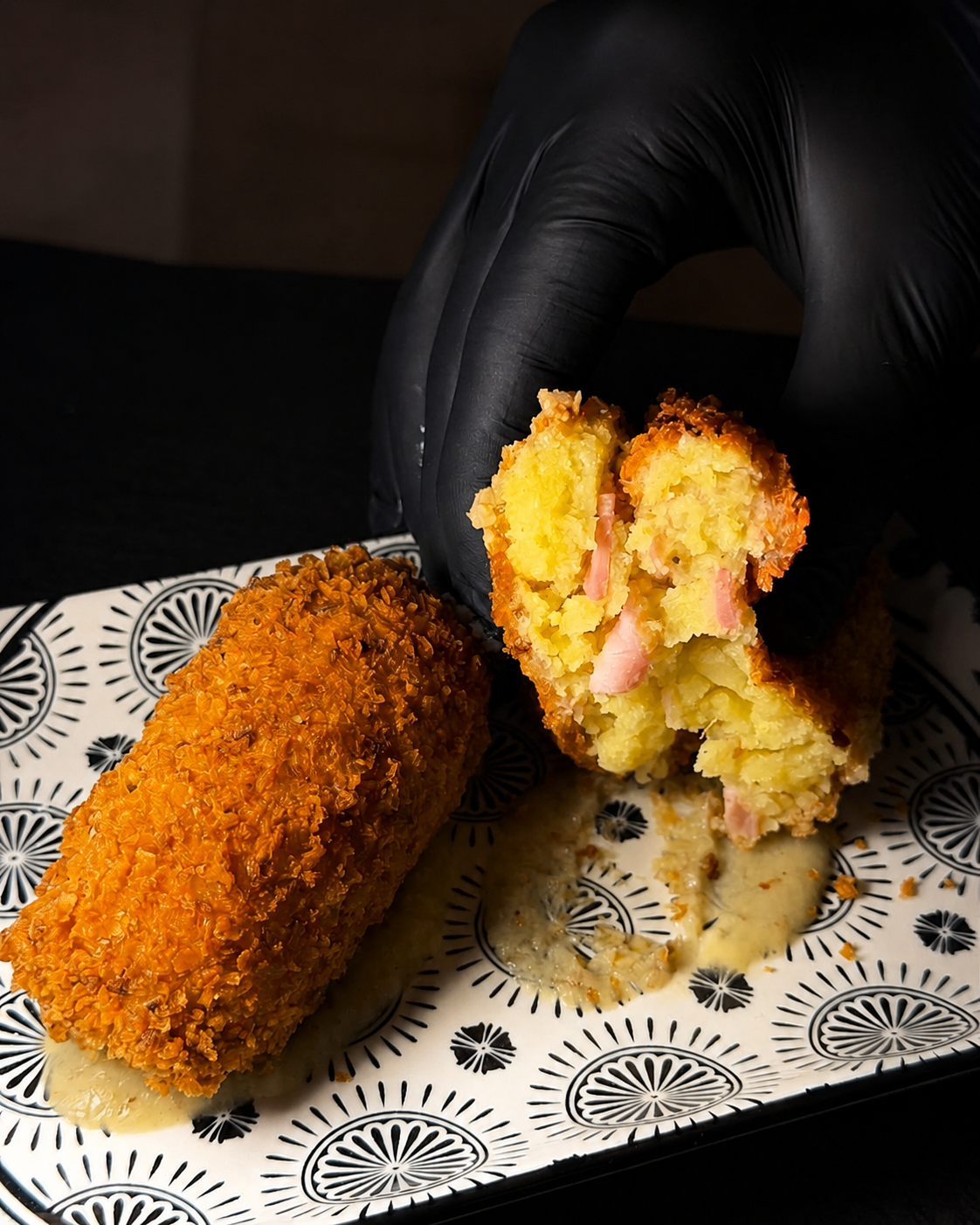 Mano guantata che regge una crocchetta dorata fritta divisa a metà su un piatto decorato, con una crocchetta intera accanto.