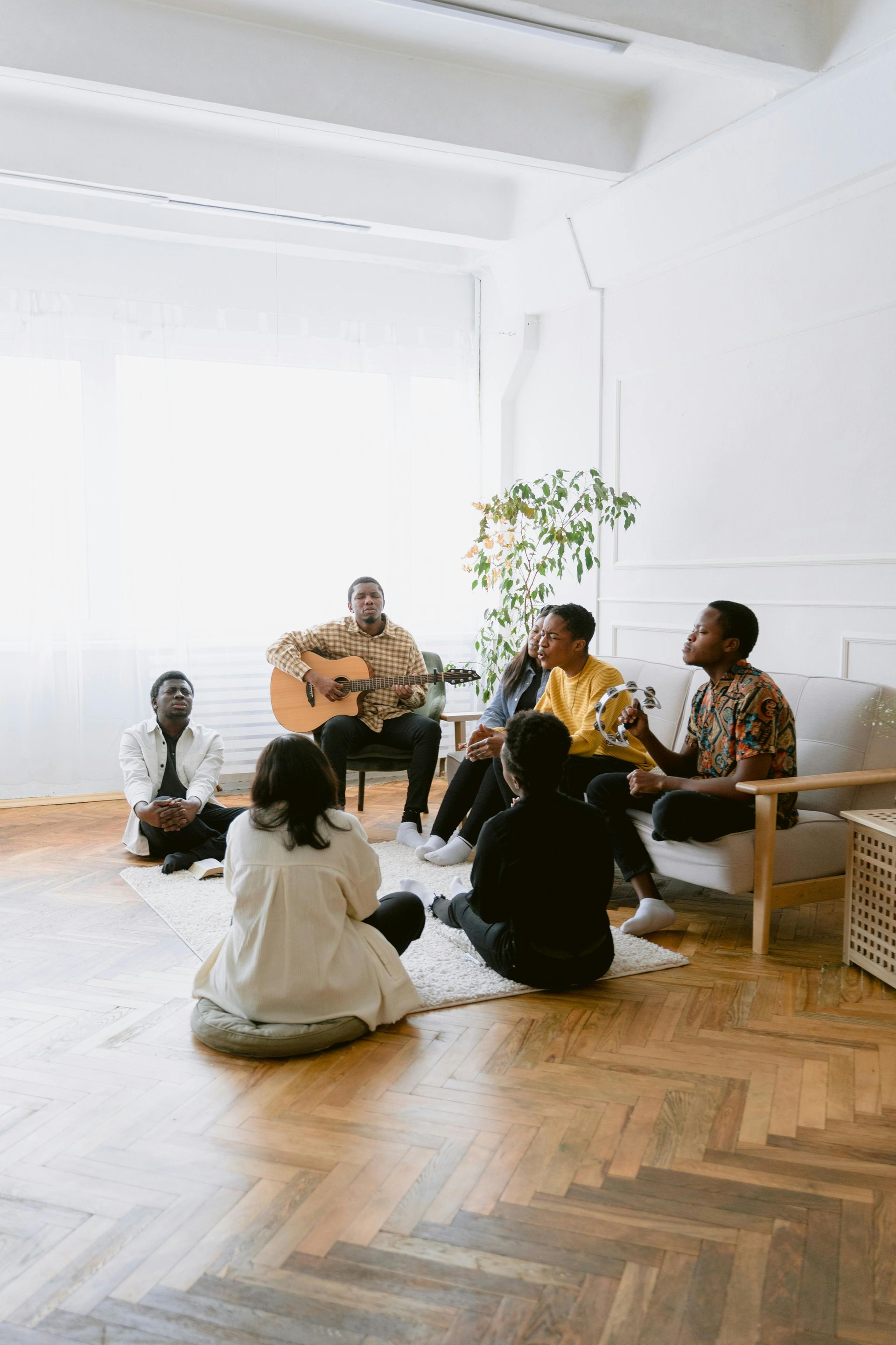 People seated in a circle, one playing guitar, in a bright minimalist room with flowers.