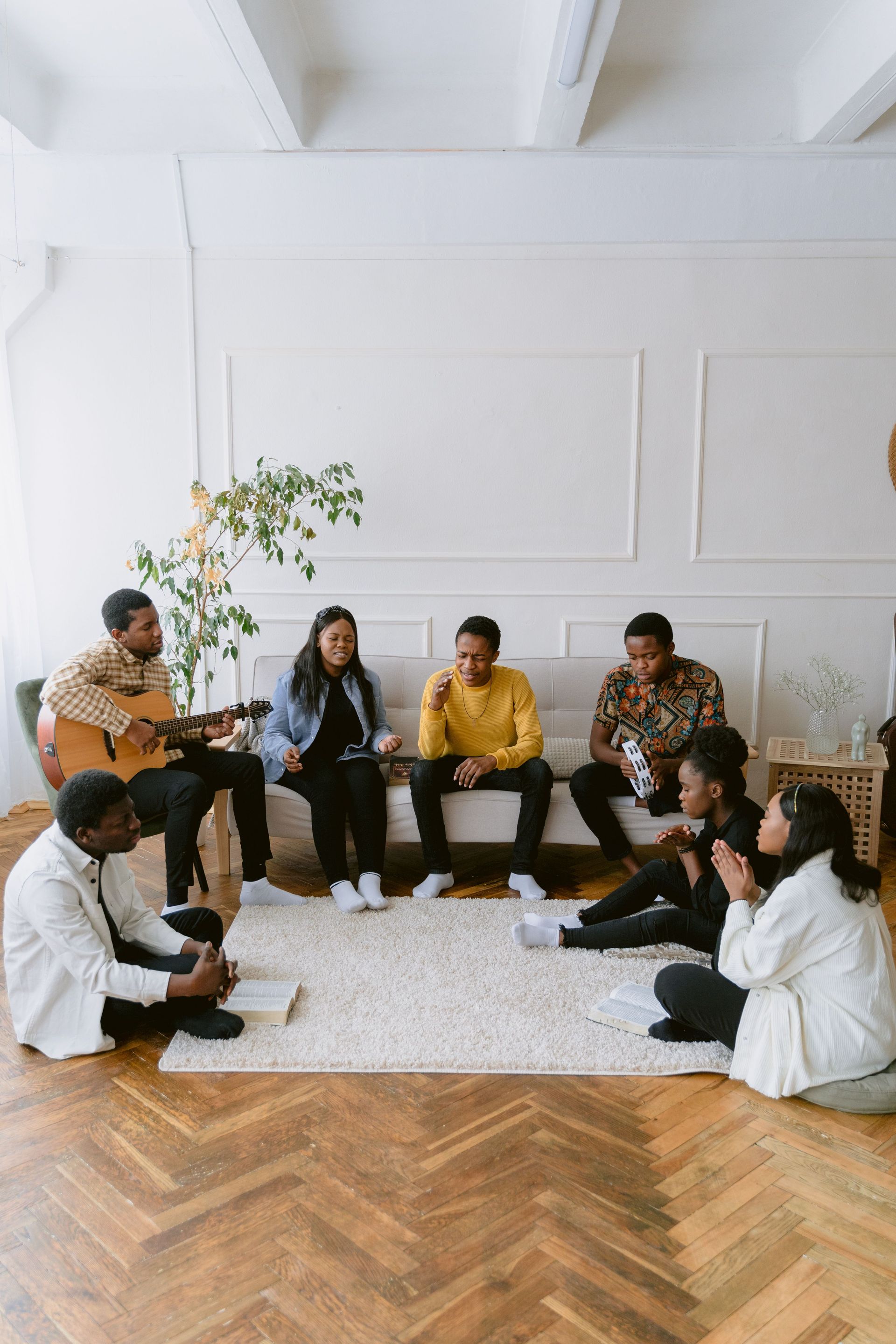 People seated in a circle in a bright room, chatting and playing guitar on a rug.office