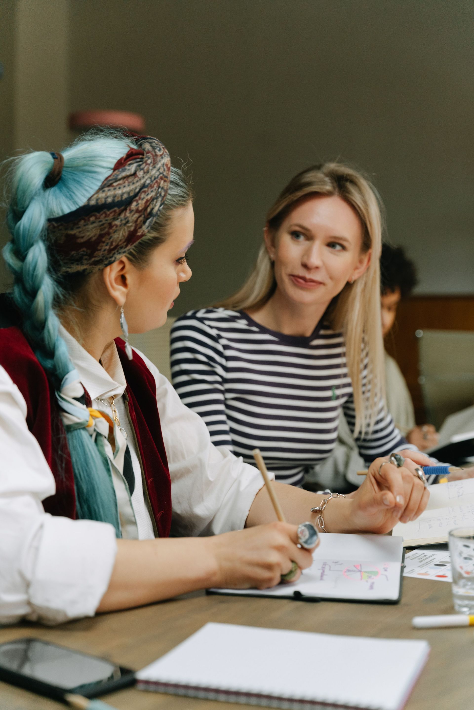 Two women in a meeting, one speaking while the other listens across a table with papers and notebooks.
