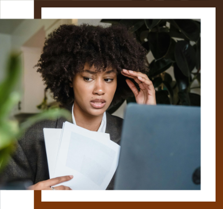 Woman looking stressed while working on a laptop, holding papers.