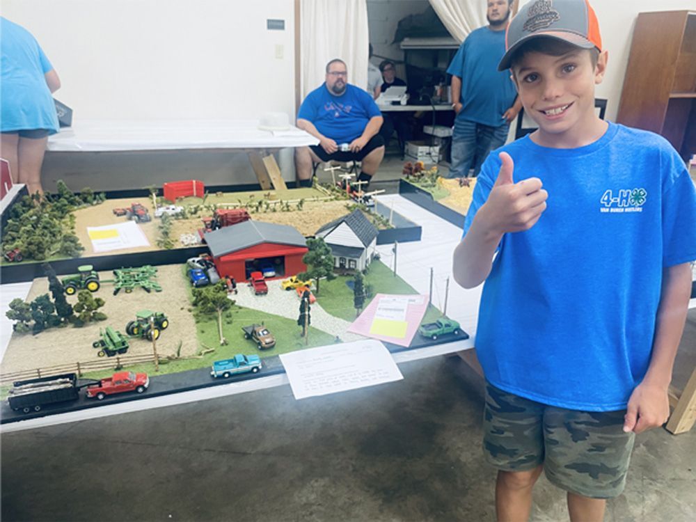 Boy in blue shirt gives thumbs-up beside a detailed model display on a table, indoors.