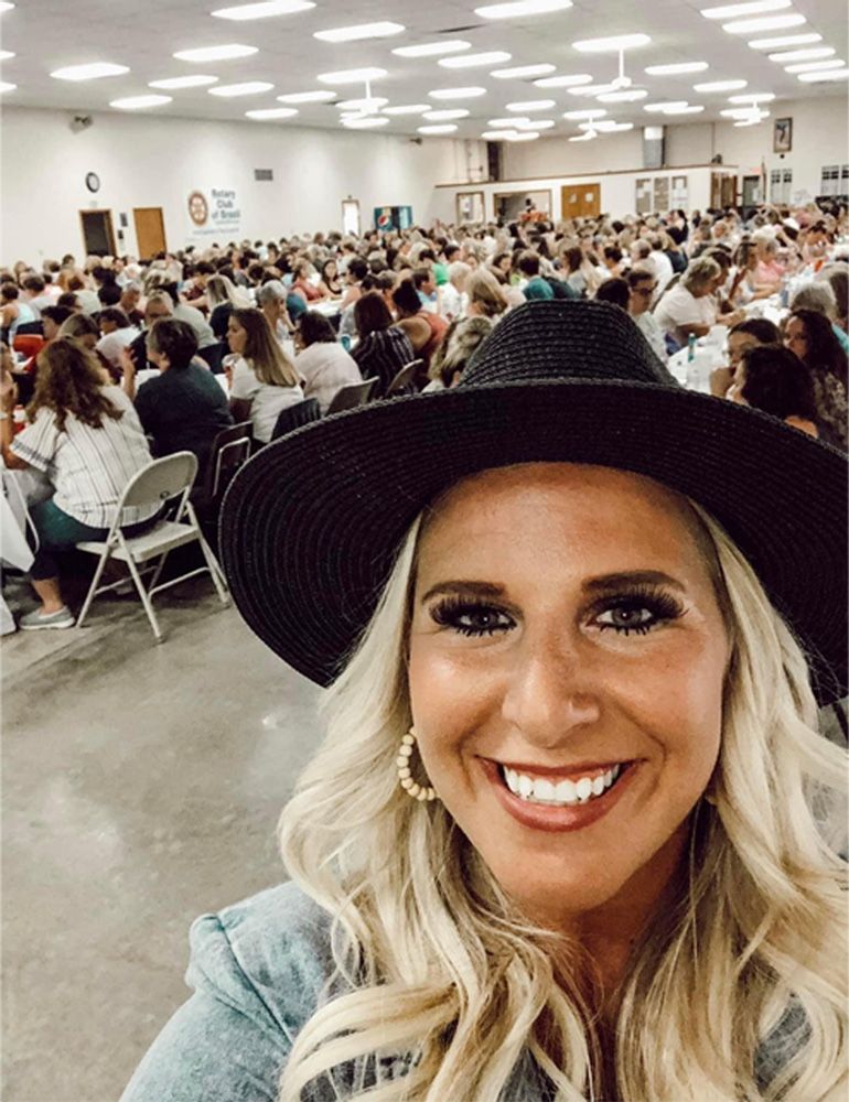 Smiling woman in a black hat taking a selfie in a crowded banquet hall.