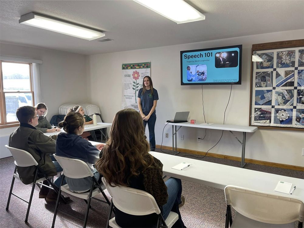Presenter giving a lesson in a classroom with students seated and a screen displaying “Lesson 101”