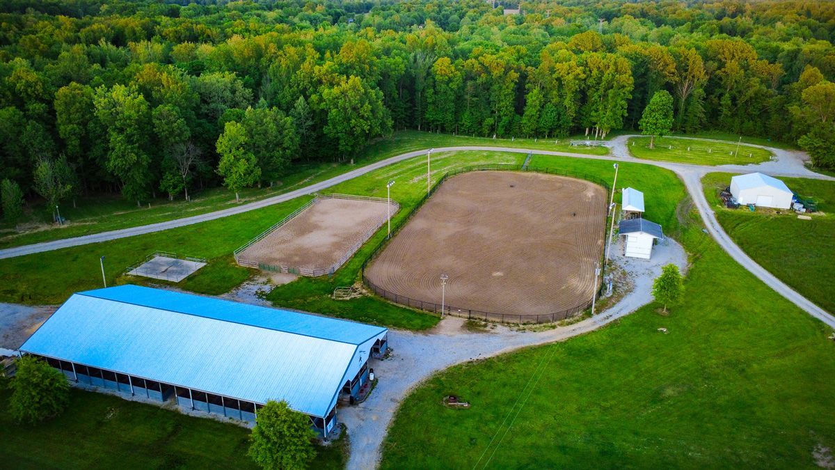 Aerial view of a farm with a blue-roofed barn, two fields, and winding gravel roads beside a wooded area