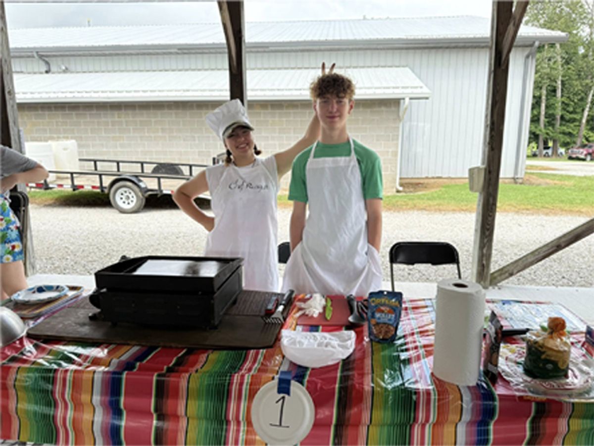Two people behind a decorated table at a booth, with a grill, snacks, and a “1” sign under a pavilion.