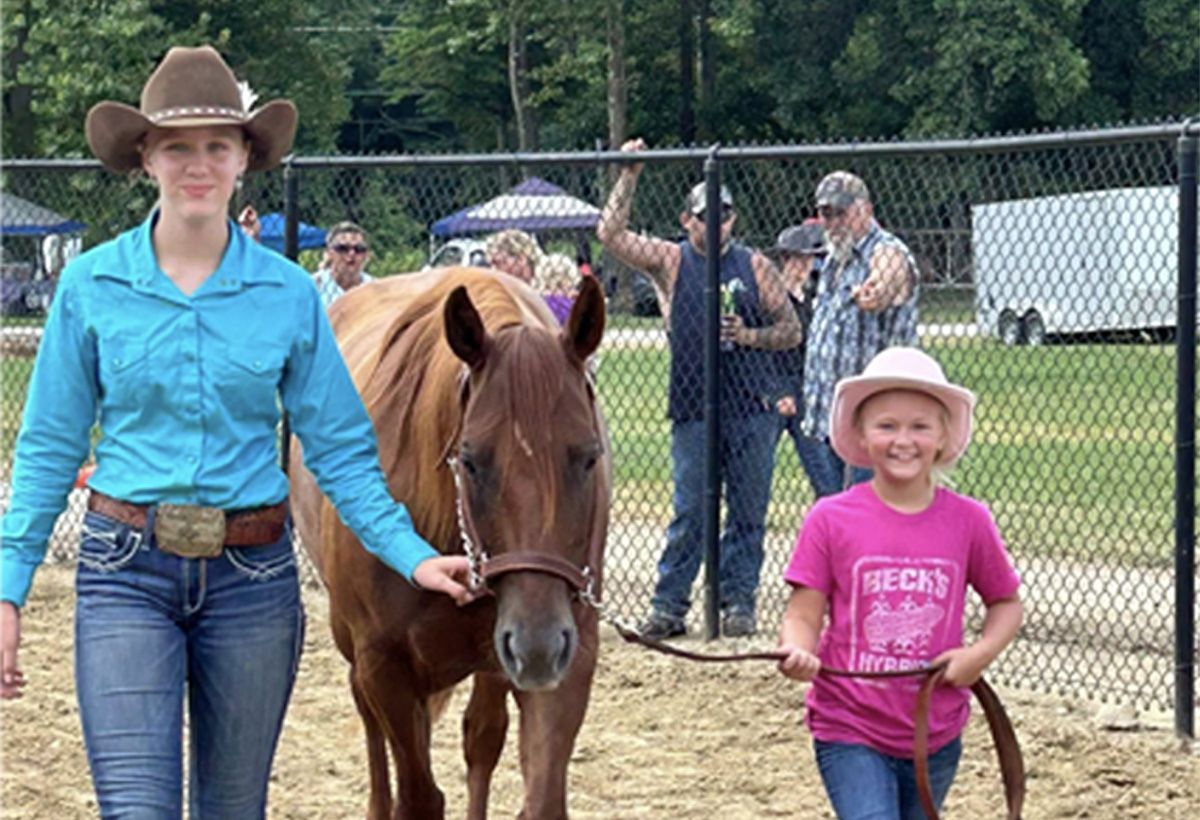 Cowgirl leading a pony beside a smiling child in pink at an outdoor fairground.