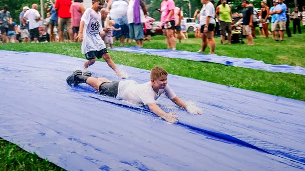 Person sliding headfirst on a wet blue tarp at an outdoor event, with spectators in the background