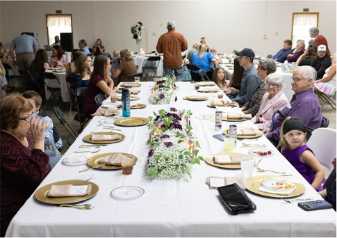 Community banquet with long tables set for a meal, people seated and chatting in a bright dining hall.