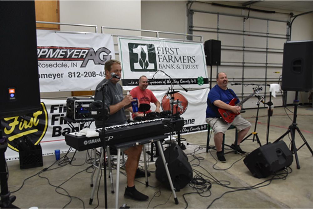 Three musicians perform indoors with keyboards, guitar, and microphone stands at a small venue