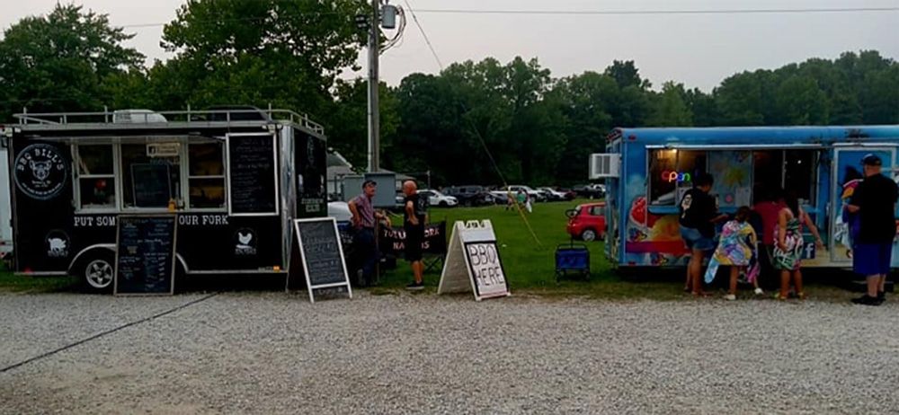 Two food trucks at an outdoor event, with people gathered around and trees in the background.