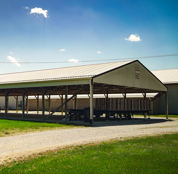 Open-sided barn with metal roof on a sunny grassy farm, with a gravel path and blue sky