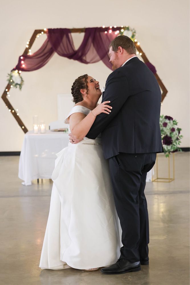 Bride and groom dancing at their wedding reception, standing beneath purple drapery and string lights